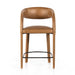 Counter stool with tan upholstered seat and back, curved arms, wood legs, black footrest.