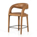 Counter stool angled view with tan upholstered seat, curved back, and black metal footrest ring.