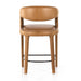 Counter stool front view with tan upholstered seat and back, curved arms, wood legs, black footrest.