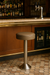 Tufted round modern bar stool featuring performance upholstery seat and silver metal stand in front of a bar with bottles and a faucet in the background.