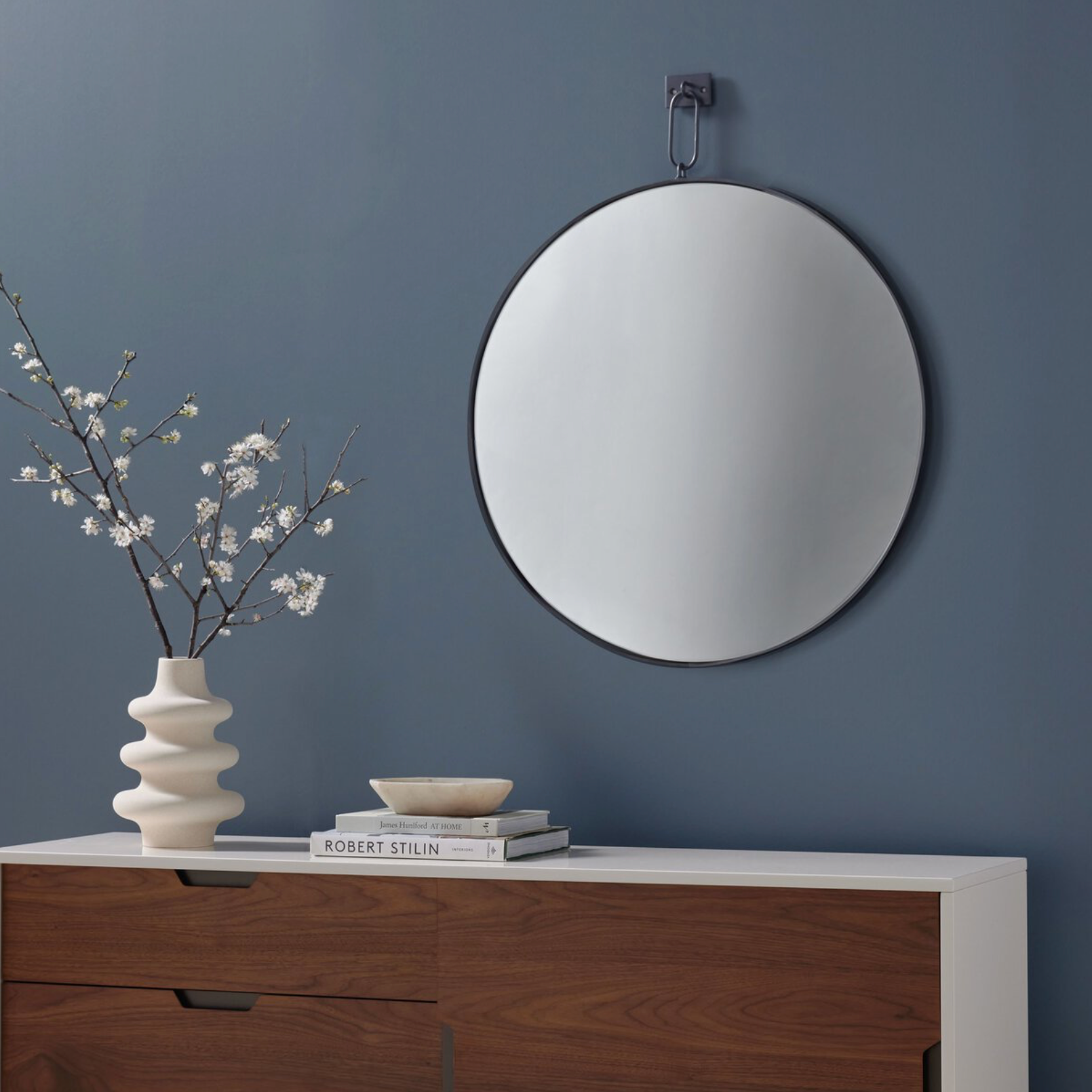 Vina square wall mirror featuring a unique vertical suspension bar in a hand-applied antiqued iron finish on a blue wall above a wooden cabinet with decorative items