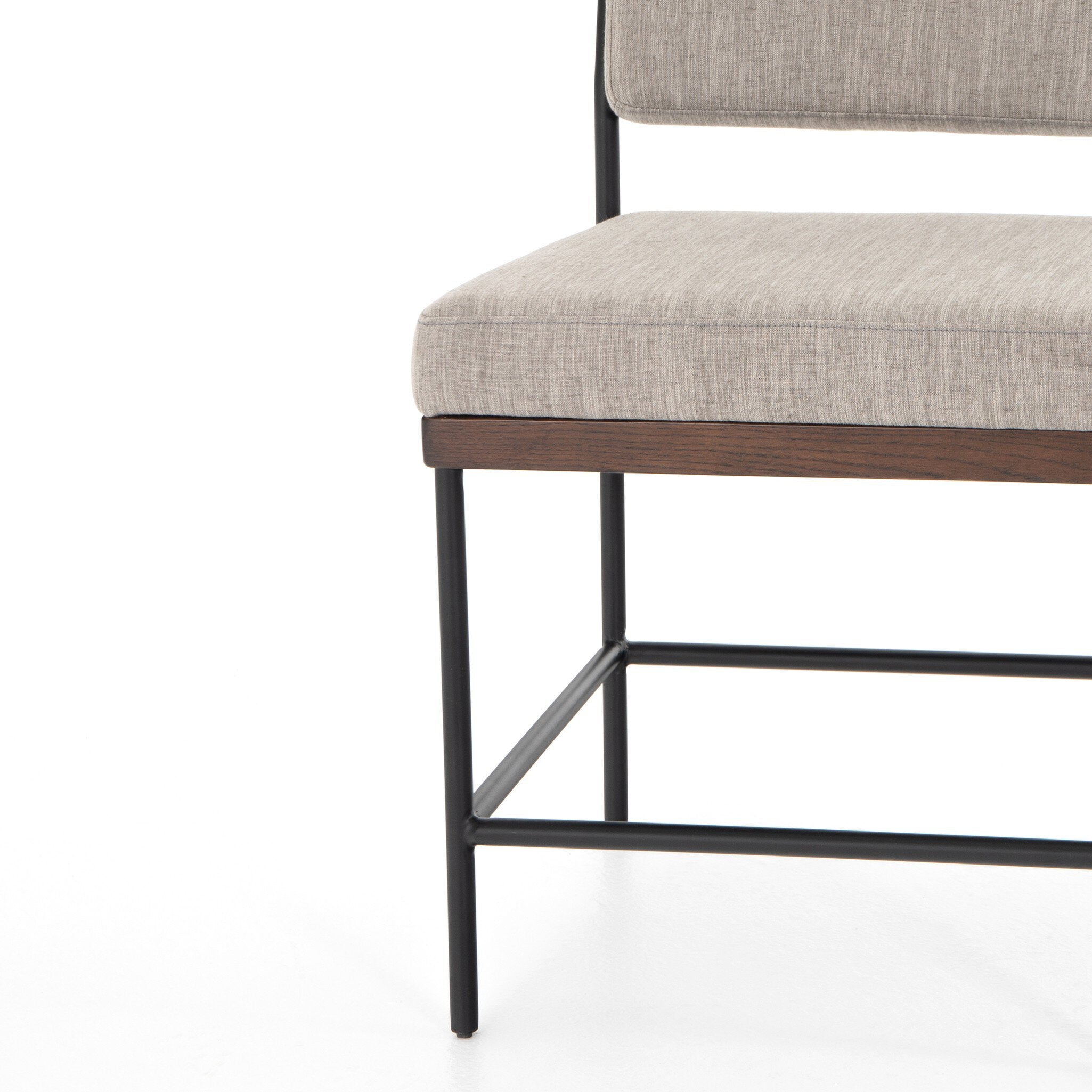 Chair with beige cushioned seat and wooden frame on a white background. Corner chair detail Benton Dining Chair in Savile Flannel performance fabric featuring a slim iron frame and wood bar.