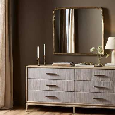 Gray dresser with a Loire mantel mirror with an arched silhouette and hand-applied antiqued gold leaf aluminum frame above it in a room with brown walls and a curtain