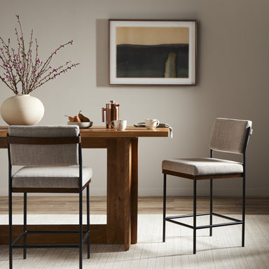 Dining area with wooden table and two bar stools against a neutral wall with a painting. Benton Counter Stool in Savile Flannel performance fabric with slim iron frame and nettlewood detailing.