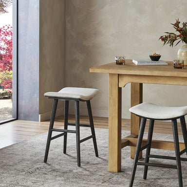Two bar stools in front of a wooden table with a neutral background. Union Bar Stool in Essence Natural fabric with a light carbon-finished oak saddle-style seat.