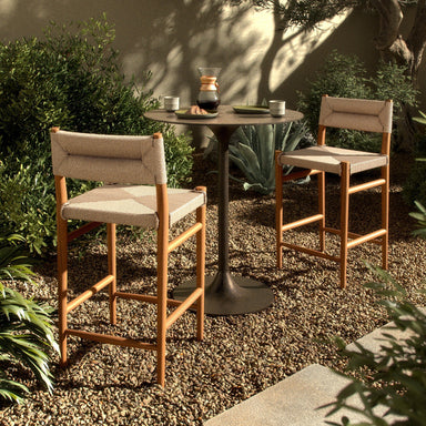 Two Lomas Counter Stool in Vintage White woven wicker with a tapered natural teak frame around a small round table in an outdoor setting.