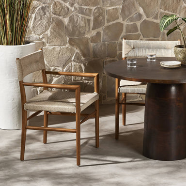 Dining area with Lomas Outdoor Dining Armchair in Vintage White woven wicker with a tapered natural teak frame and a round table against a stone wall