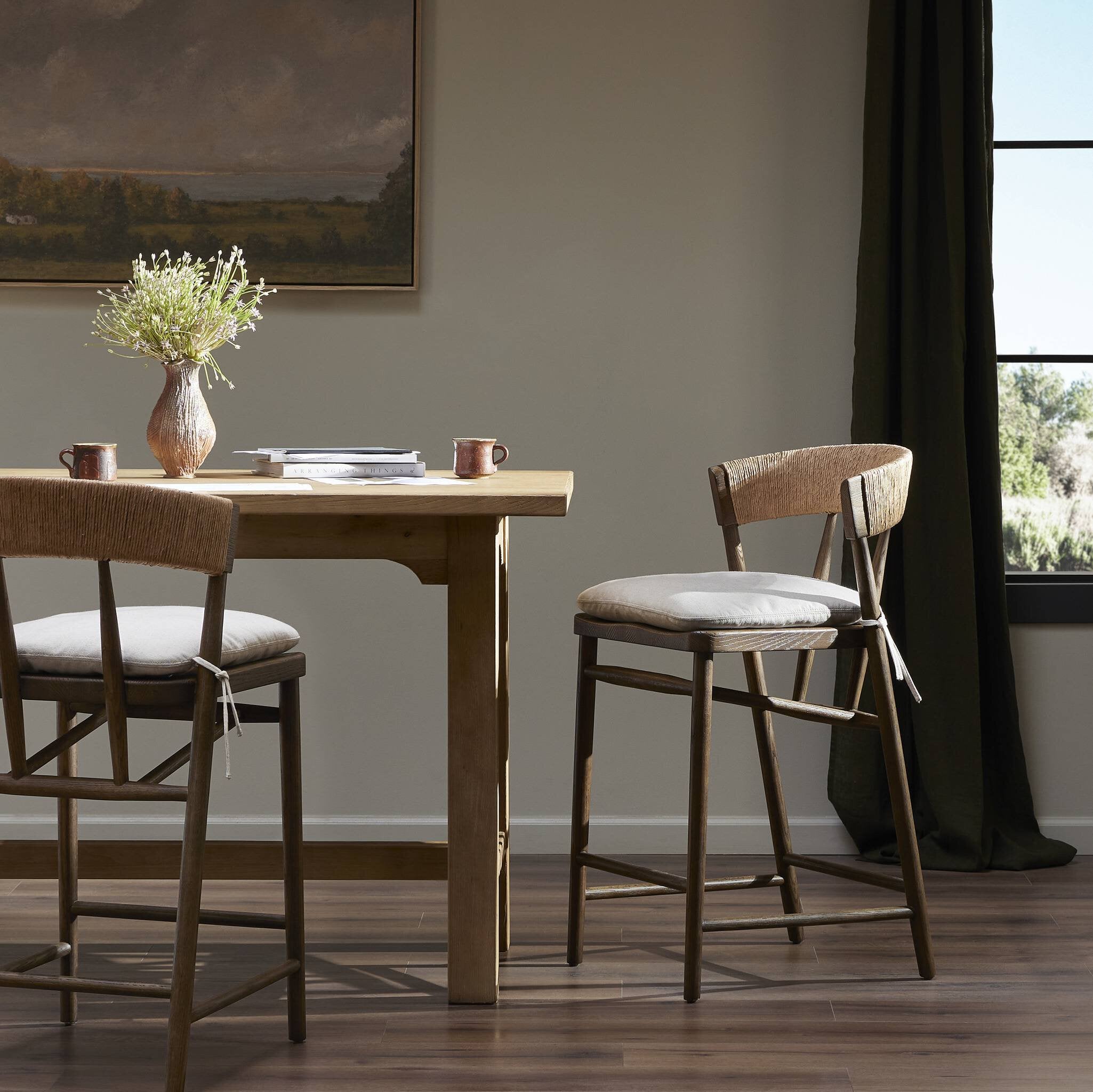 Dining area with wooden table and Buxton Bar Counter Stool in solid ash with a hand-wrapped paper rush backrest in a room with a painting on the wall.