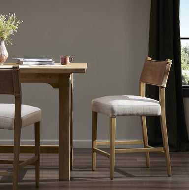 Wooden dining table with Contract grade Ferris beige counter stool with a wood frame and fabric seat, designed for high-performance fabric with a leather backing in a room with a window and plant.