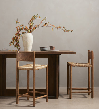 Wooden Erie Bar Counter Table in Smoked Oak with a chunky pedestal base and modern rustic designwith two chairs and a vase of flowers against a plain wall.