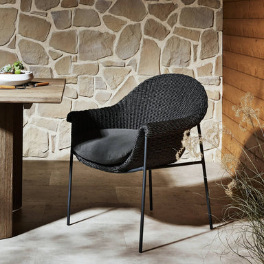 Suerte wicker outdoor dining chair in arashi ink with faux hyacinth weave and iron base with dark cushions against a stone wall
