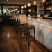 Theo bar stool in bar setting, beige woven back with dark round seat and wood frame