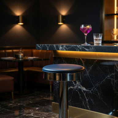 Hawthorne Bolted Floor-Mounted Counter Stool with a smooth round navy blue vinyl seat and a polished chrome pedestal base in front of a marble bar counter with drinks in a dimly lit bar