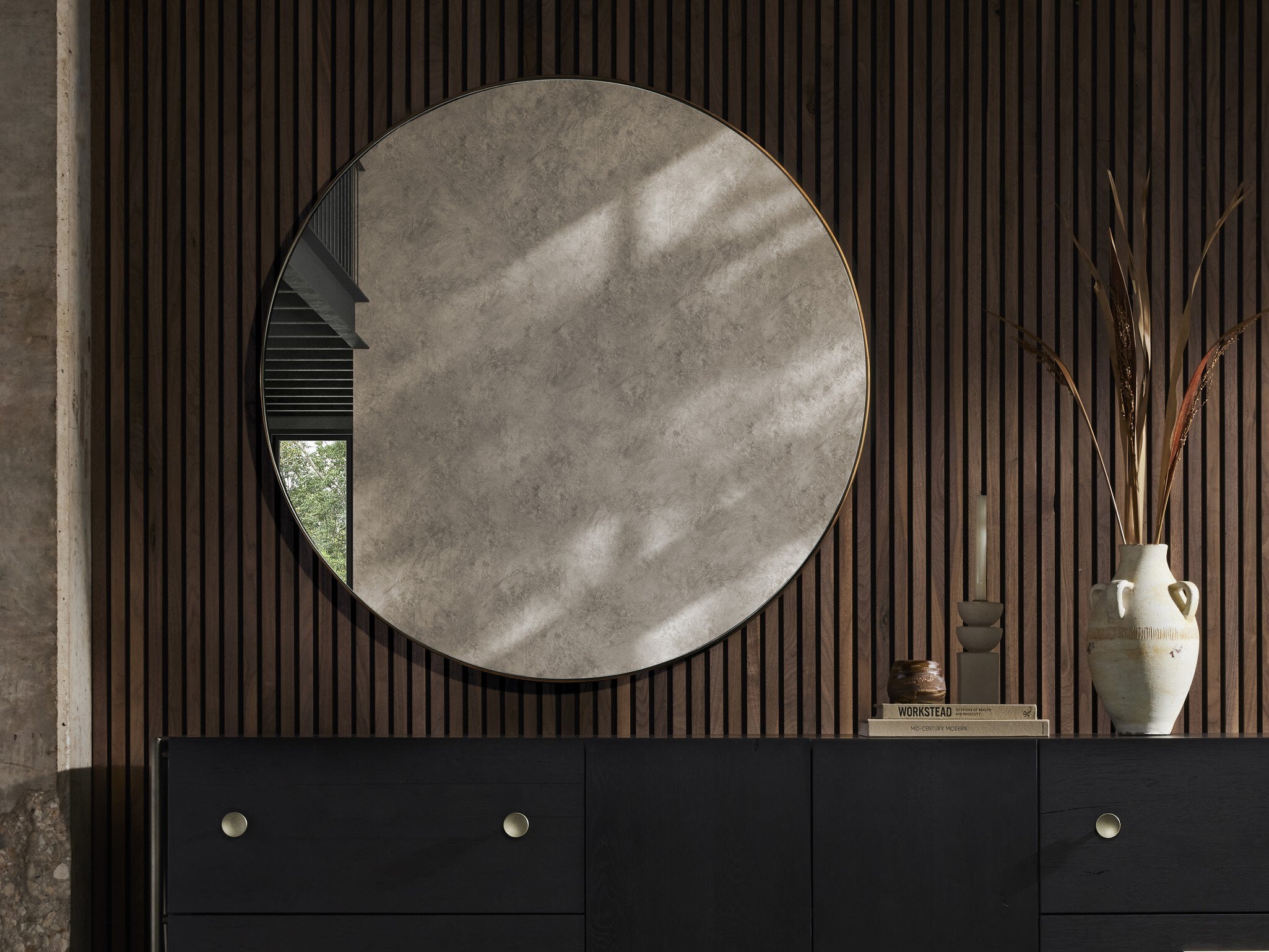 Georgina 47-inch oversized round mirror with a slender polished brass aluminum frame on a wooden wall above a black dresser with decorative items