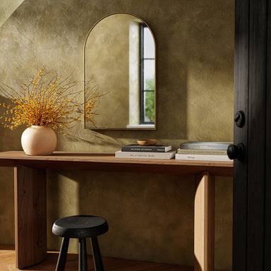 Wooden console table with Georgina 21-inch arched wall mirror with a slender polished brass-finished aluminum frame, books, and vase against a textured wall