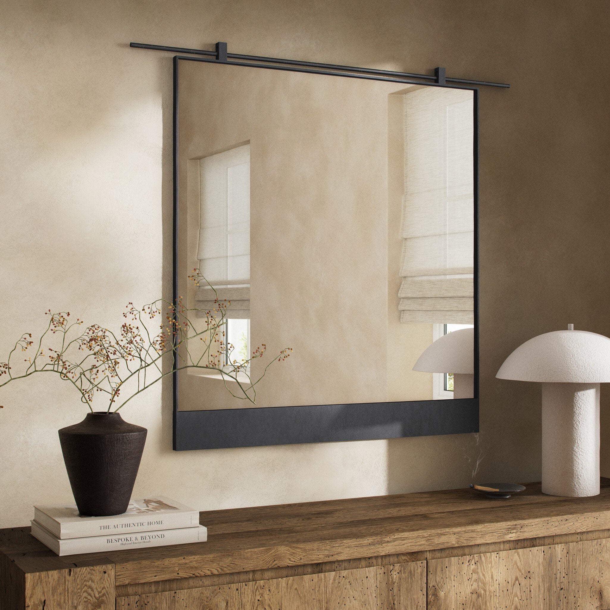 Chico square wall mirror featuring a unique vertical suspension bar in a hand-applied antiqued iron finish with black frame on a beige wall above a wooden console table with decorative items