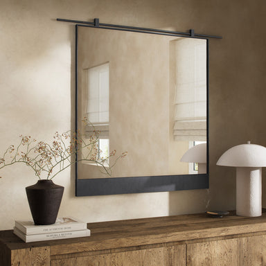 Chico square wall mirror featuring a unique vertical suspension bar in a hand-applied antiqued iron finish with black frame on a beige wall above a wooden console table with decorative items