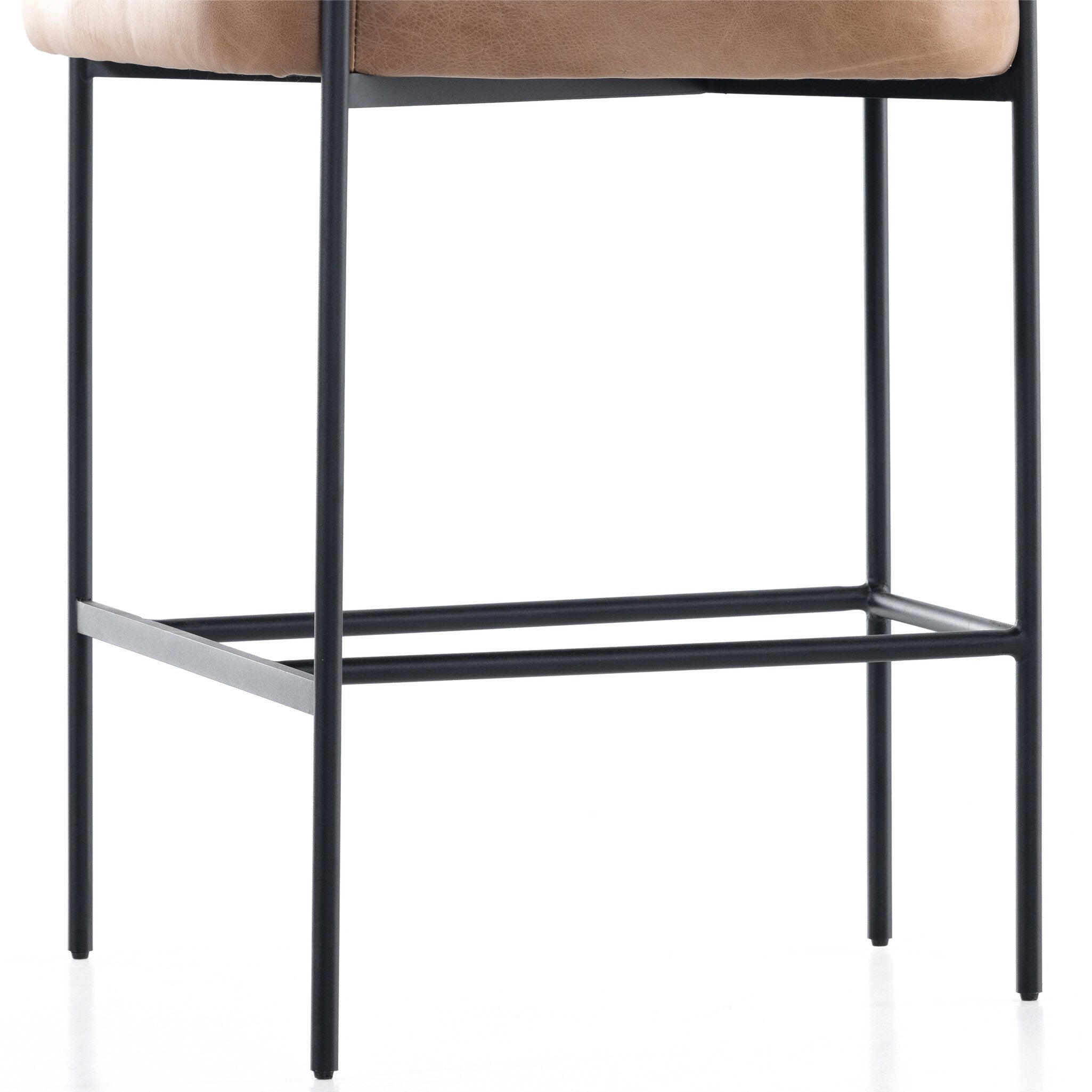 Bar stool with brown seat and black metal frame on a white background. Black leg detail Carrie Bar Stool in Chaps Saddle performance fabric with black-finished iron framing and yoke back detailing.