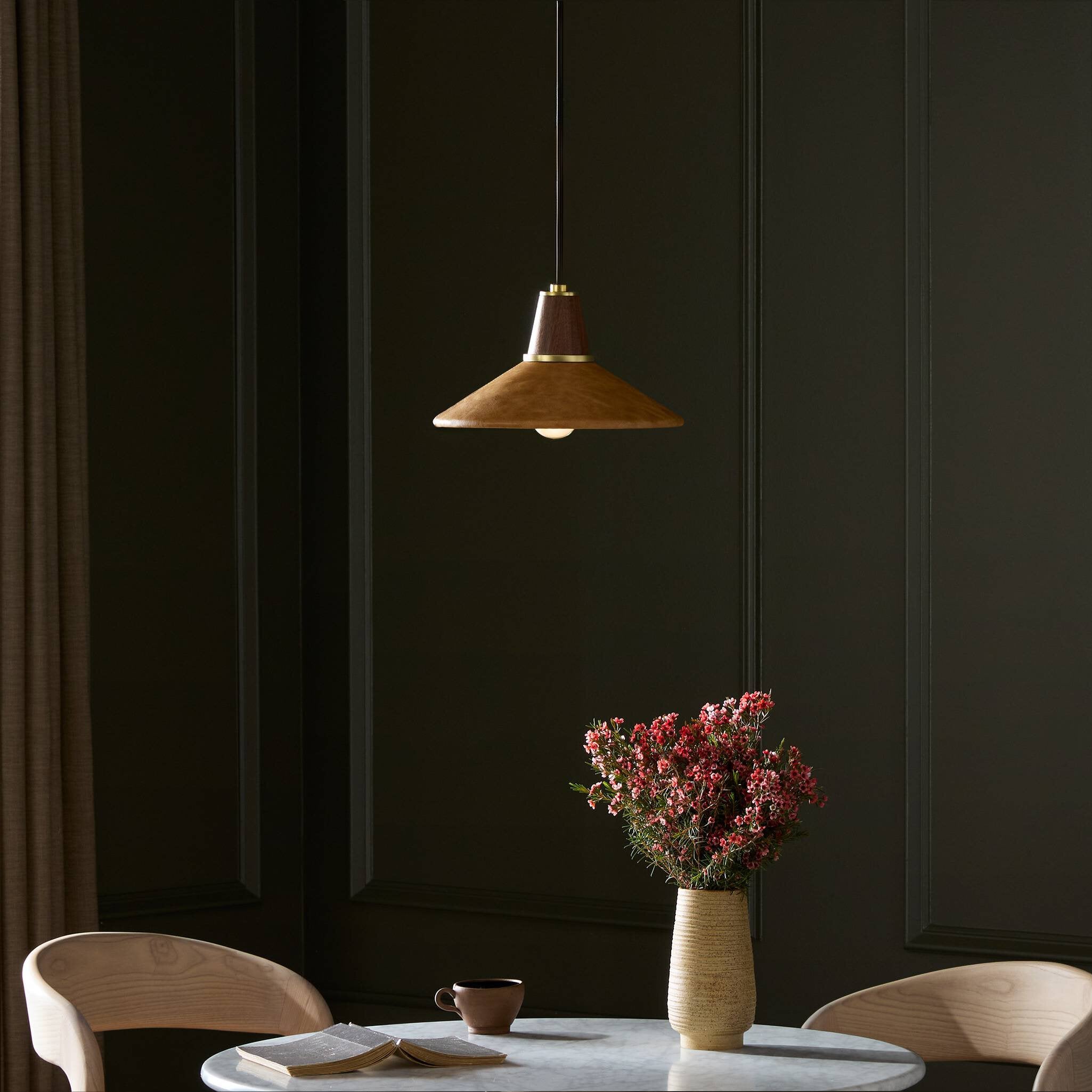 Modern interior with a mid century Cullen pendant light with a camel leather shade and natural walnut finial, table, chairs, and a vase with flowers against a dark wall