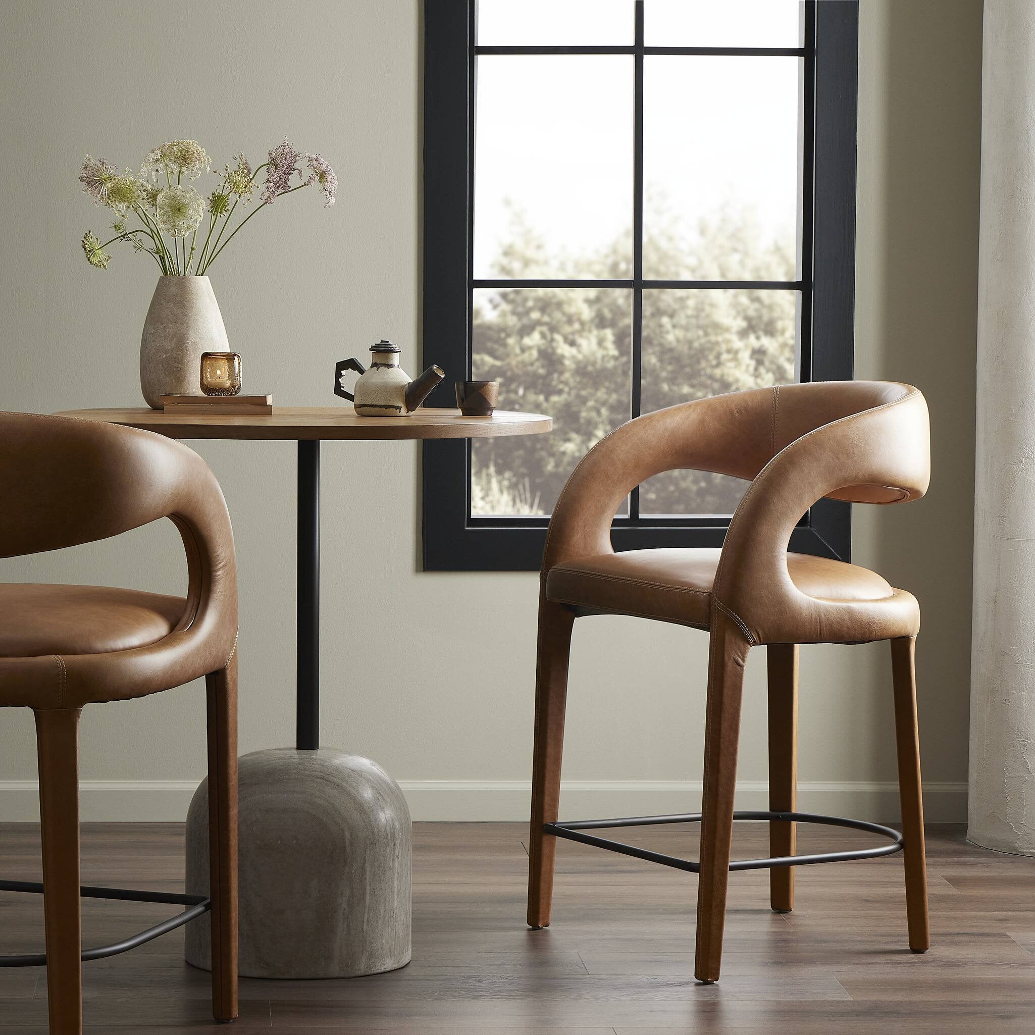 Counter stool with tan leather-look upholstery and curved arms, on wood legs with black footrest.