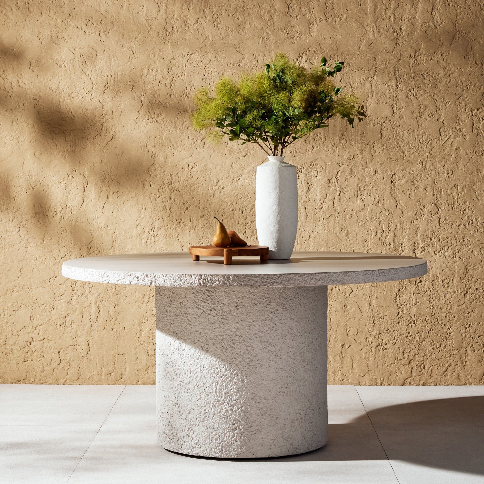 Modern industrial monolithic round outdoor dining table made of solid white concrete with a vase and decorative items against a beige wall.