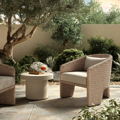 An outdoor chair with a barrel-shaped back, a white cushion, and three legs, made from all-weather wicker with beige cushions and a small table on a patio with greenery.